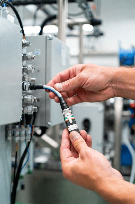 Close-up of technician installing high-flex modular sensor actuator cable with M12 connector into junction box on factory machinery, demonstrating fast and error-free wiring transition