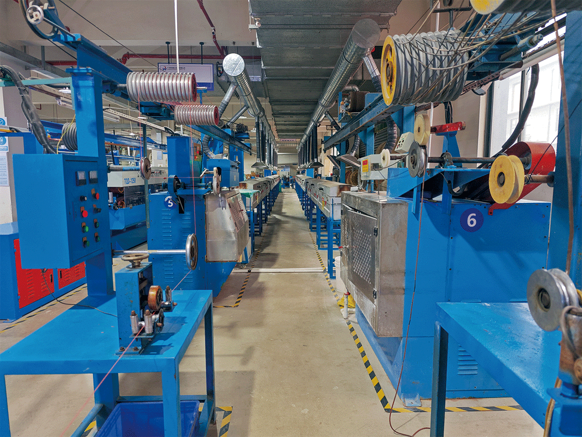 Interior view of a HULK cable production facility showing rows of blue automated machines (labeled 5, 6) engaged in stranding, shielding, or jacketing processes; overhead metal conduits and wire spools indicate integrated material flow, while floor markings and control panels reflect strict operational standards for consistent, high-volume cable manufacturing.