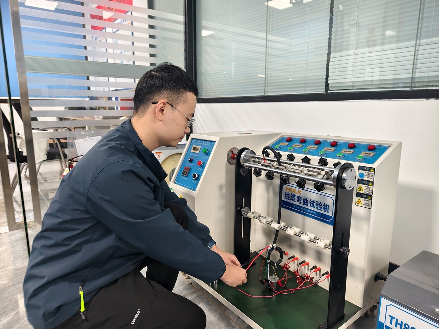 An engineer operating a HULK Cable Bending Test Machine in a lab, with multiple cables undergoing repeated flexing cycles; the setup includes real-time monitoring probes and data logging to evaluate conductor fatigue, insulation degradation, and shielding integrity under dynamic stress for R&D validation.