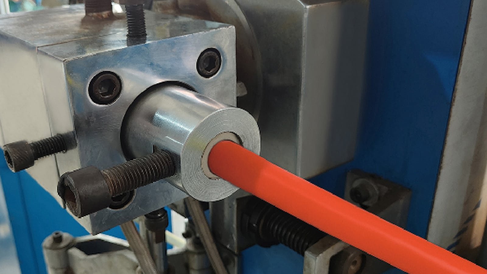 A close-up of an orange HULK high-flex cable being extruded through a precision die in a manufacturing line; the setup includes real-time diameter monitoring and tension control to ensure consistent insulation thickness, concentricity, and surface finish during high-speed production for robotics or automotive applications