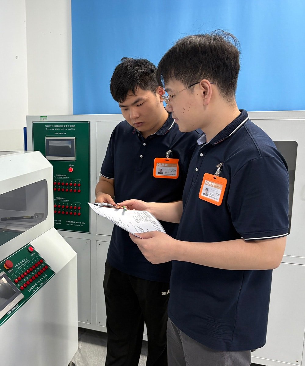 HULK engineers reviewing test data beside a wire drag chain testing machine in a quality assurance lab, demonstrating rigorous validation of cable durability and performance under simulated industrial stress conditions.