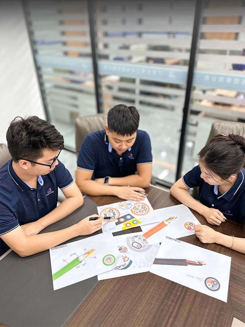 Three HULK engineers collaborating around a table covered with cable cross-section design drafts, discussing conductor layout, insulation thickness, and shielding configurations — reflecting the brand’s commitment to co-developing tailored cable solutions that meet unique application requirements from the earliest design phase.