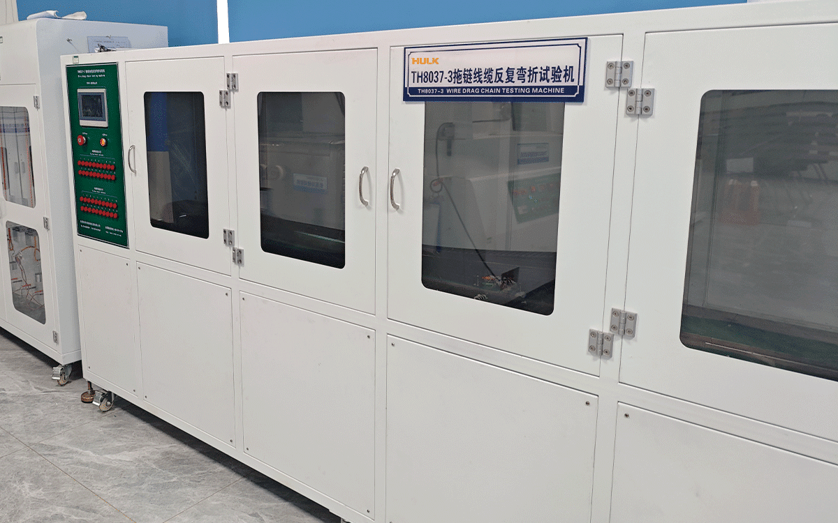 A HULK TH8037-3 Wire Drag Chain Testing Machine housed in a clean laboratory environment; this specialized white equipment with a green control panel and viewing windows performs rigorous repetitive bending tests to validate the mechanical durability and service life of HULK's high-flex drag chain cables under dynamic motion conditions.