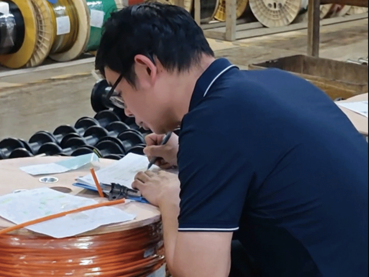 A HULK quality inspector conducting final outgoing checks at the production site, writing verification data on a clipboard resting on a large orange cable reel; surrounded by stacked black reels in the warehouse, this scene highlights the rigorous manual oversight and documentation process ensuring every shipped product meets specifications before delivery.