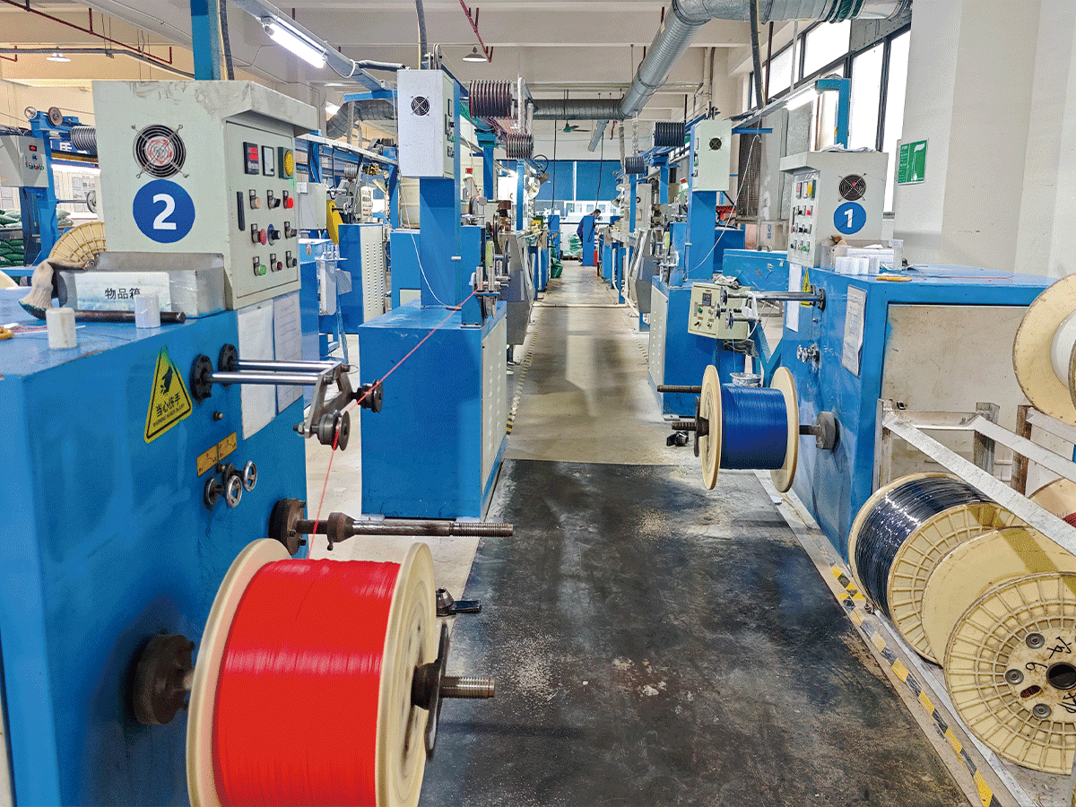 HULK factory production floor featuring a long aisle of numbered blue cable manufacturing machines (labeled '1' and '2'); large red and blue cable reels are actively rotating in the foreground, while a worker monitors the line in the distance, illustrating high-capacity parallel processing and continuous operation capabilities.