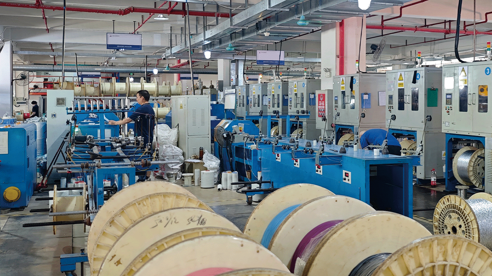 HULK factory braided shield production area featuring automated blue machines under labeled zones 'BRAIDED REGION' and 'BRAIDER SHIELD AREA'; workers operate equipment while cable reels are staged in foreground, showcasing in-house manufacturing of EMI-resistant shielding layers for high-performance industrial cables.