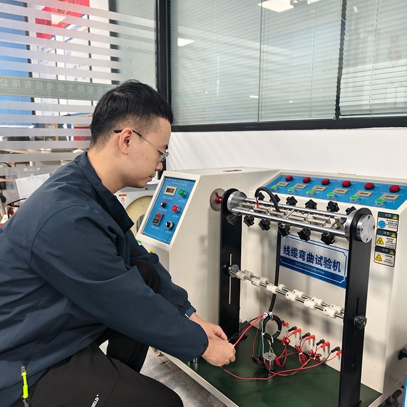 HULK engineer operating a multi-channel cable bending fatigue tester, connecting test leads to samples under repeated flexing motion; machine features digital control panel and multiple test stations — validating long-term mechanical endurance and electrical continuity of cables under simulated real-world bending cycles.
