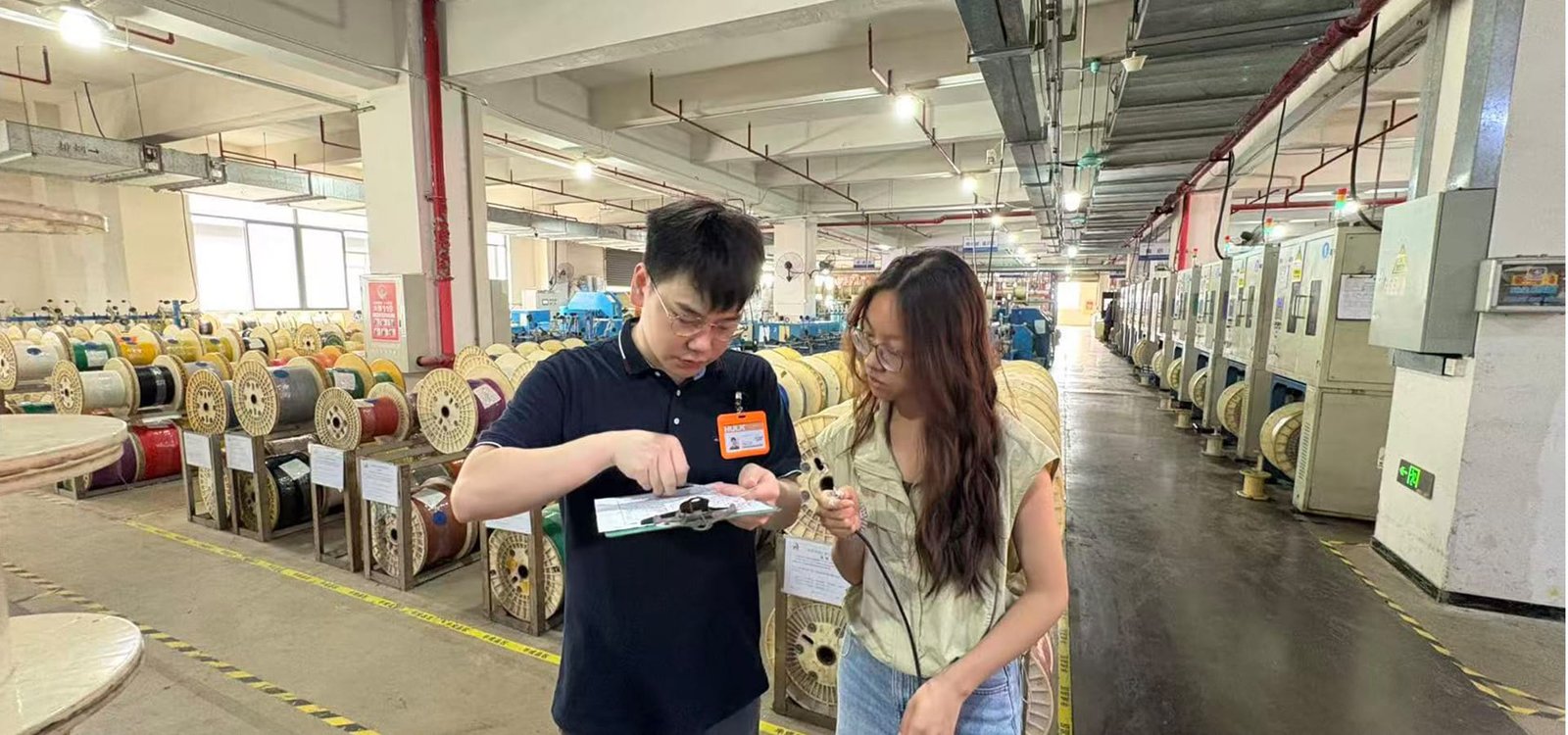 HULK factory production floor showing engineers conducting on-site quality inspection; a male technician with HULK badge reviews test data on clipboard while explaining cable sample details to a colleague, surrounded by rows of finished cable reels and manufacturing equipment, highlighting real-time quality control and technical expertise.