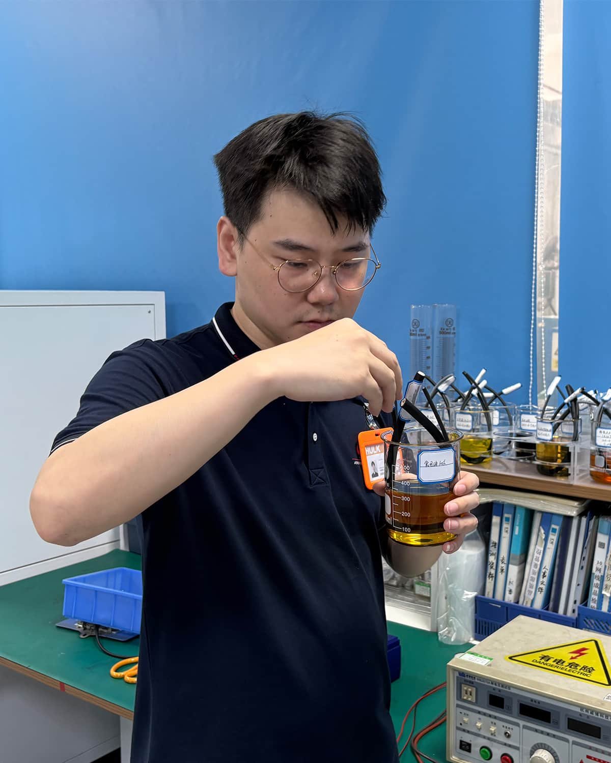 HULK R&D engineer conducting oil resistance test on cable samples submerged in amber chemical solution within a beaker, surrounded by multiple testing stations and electrical equipment — demonstrating rigorous material validation processes to ensure cable durability and jacket integrity in harsh industrial environments.