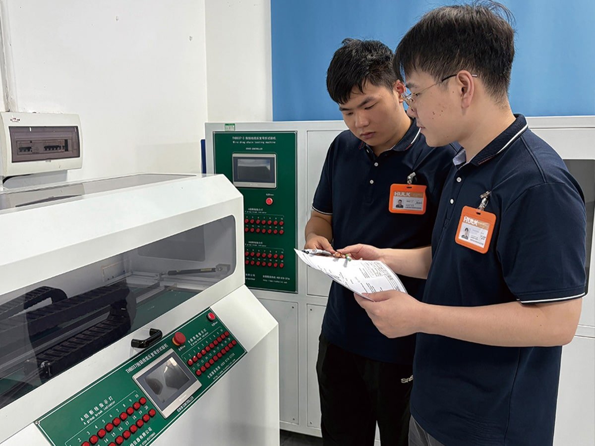 Two HULK technicians collaborating at a testing station, reviewing data on a clipboard next to a torsion testing machine to ensure accurate recording and quality compliance.