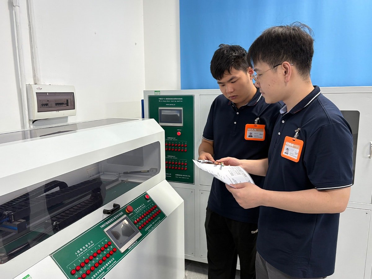 Two HULK engineers in a laboratory reviewing test data beside the TH8037 Drag Chain Cable Repeated Bending Tester; the machine features a green control panel with red indicator buttons and a transparent test chamber, demonstrating human-supervised validation of cable fatigue resistance under simulated industrial motion cycles.