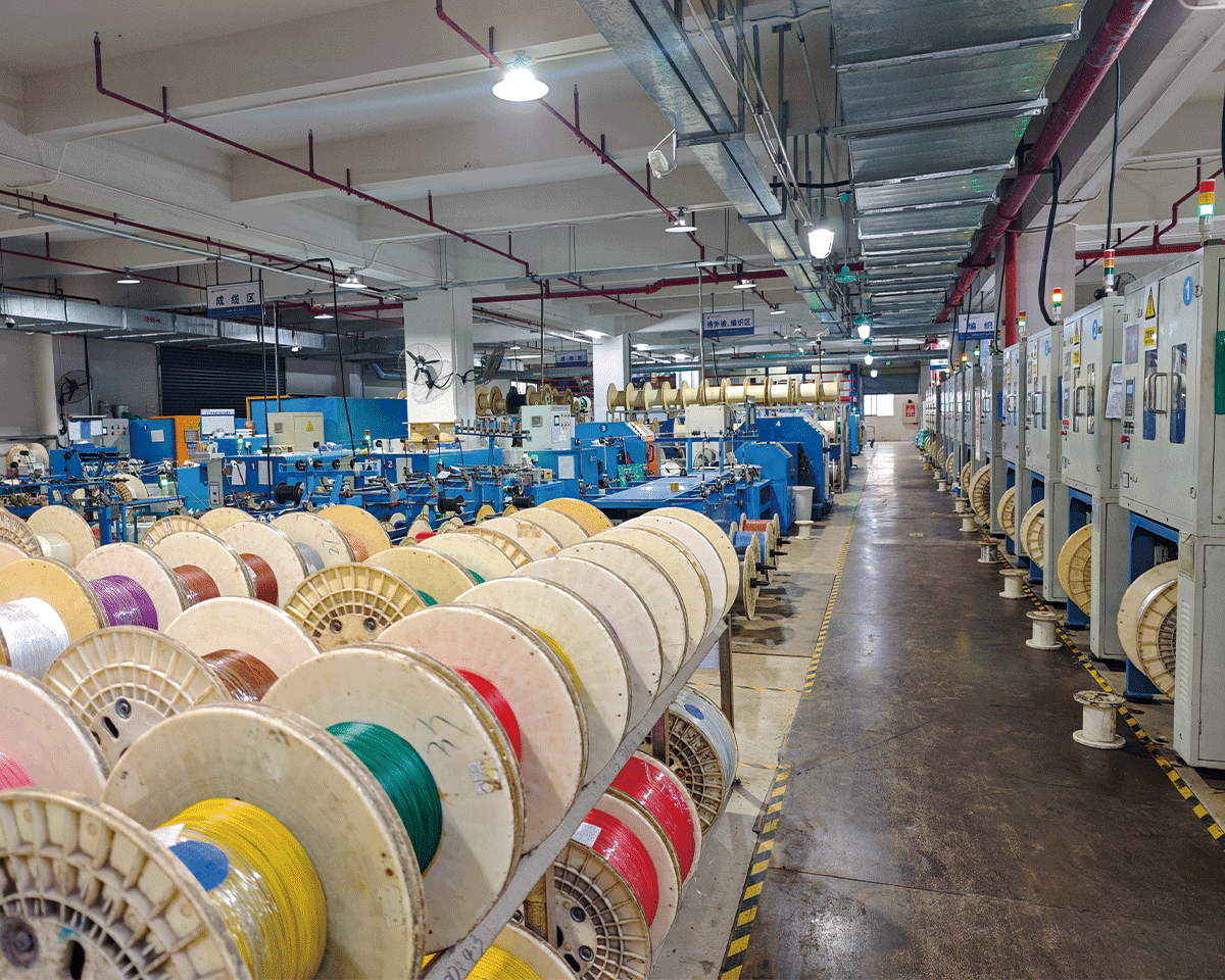 HULK factory finished goods warehouse showing rows of color-coded cable reels neatly stored on racks, with blue automated production machines and control panels visible in the background; Chinese signage indicates designated zones like 'Cabling Area' and 'Stranding Area', reflecting standardized workflow and high-capacity output for global industrial clients.