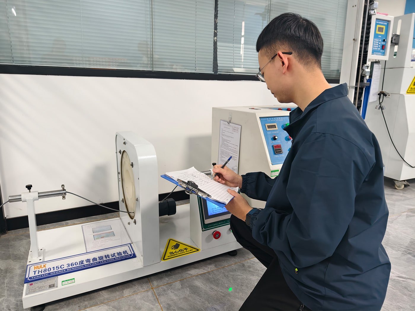 An engineer recording data on a clipboard while monitoring the HULK TH8015C 360 Degree Bending Rotary Testing Machine; the equipment features a rotating test fixture and digital control panel, validating cable durability under continuous rotational stress for applications like robotic arms and automated turntables.