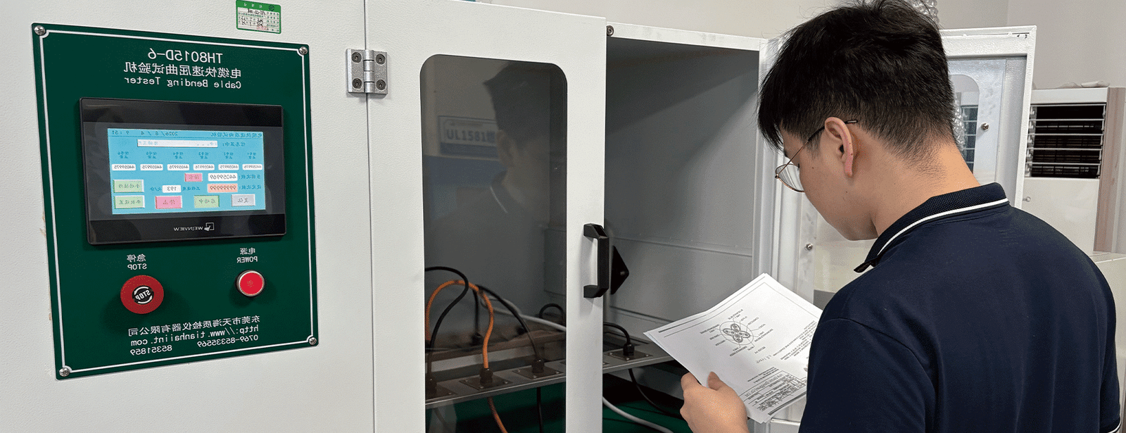 A HULK engineer reviewing a cable cross-section diagram while monitoring the TH8015D-6 Cable Bending Tester in operation; the green control panel displays real-time test data, and orange-black cables undergo high-frequency reciprocating bending inside the transparent chamber — illustrating a human-supervised validation process that ensures test parameters align precisely with client specifications.