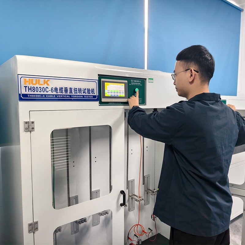 HULK TH8030C-6 Cable Vertical Torsion Tester in operation, with engineer interacting with touchscreen control panel; machine features transparent test chamber and vertical clamping fixtures — used to evaluate torsional fatigue resistance and mechanical durability of flexible cables under simulated real-world vertical motion conditions.