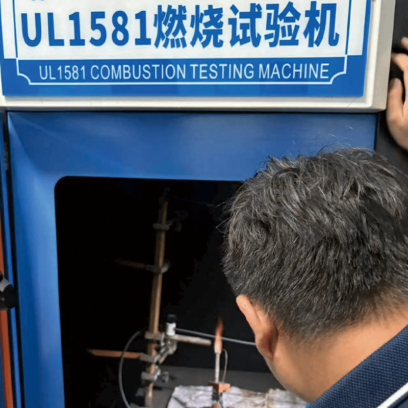 Technician observing a cable sample undergoing flame resistance testing inside a HULK UL1581 Combustion Testing Machine; the blue chamber with visible burner and clamps is used to assess how quickly flames spread and whether the material self-extinguishes, ensuring compliance with international safety standards for fire hazard mitigation.