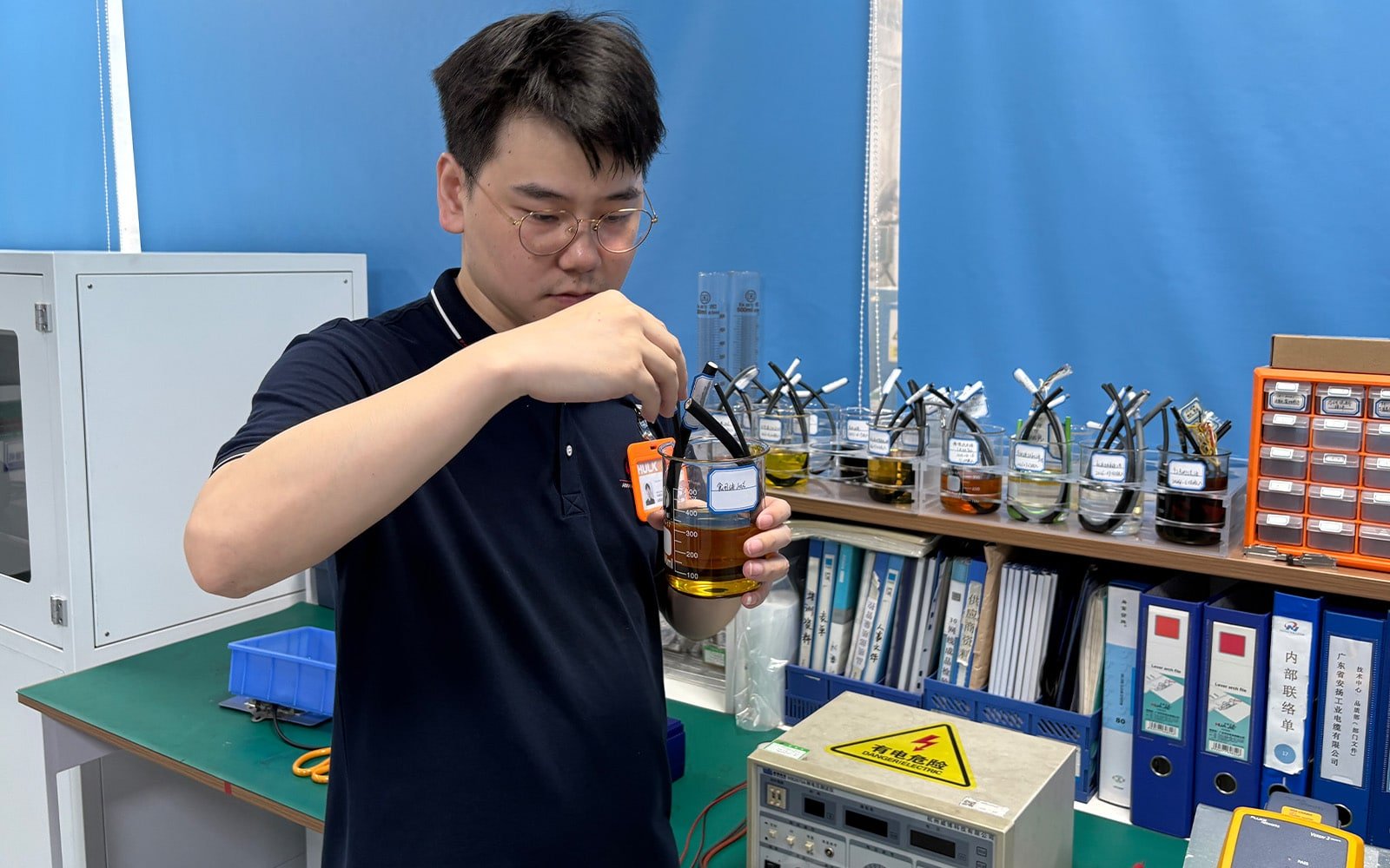 Hulk Cable lab technician inspecting cable samples immersed in chemical liquids for oil and solvent resistance testing, with multiple test beakers in the background validating material durability under harsh industrial environments