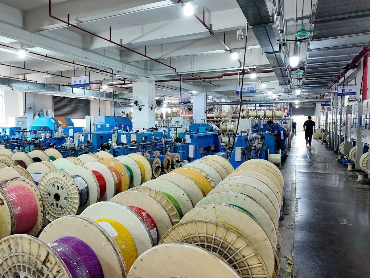 Panoramic view of Hulk Cable manufacturing workshop featuring rows of colorful cable reels in the foreground and blue production machinery in the background, with workers operating equipment under clear zone signage like 'Cabling Area' and 'Braiding Area', demonstrating large-scale production capacity and organized workflow
