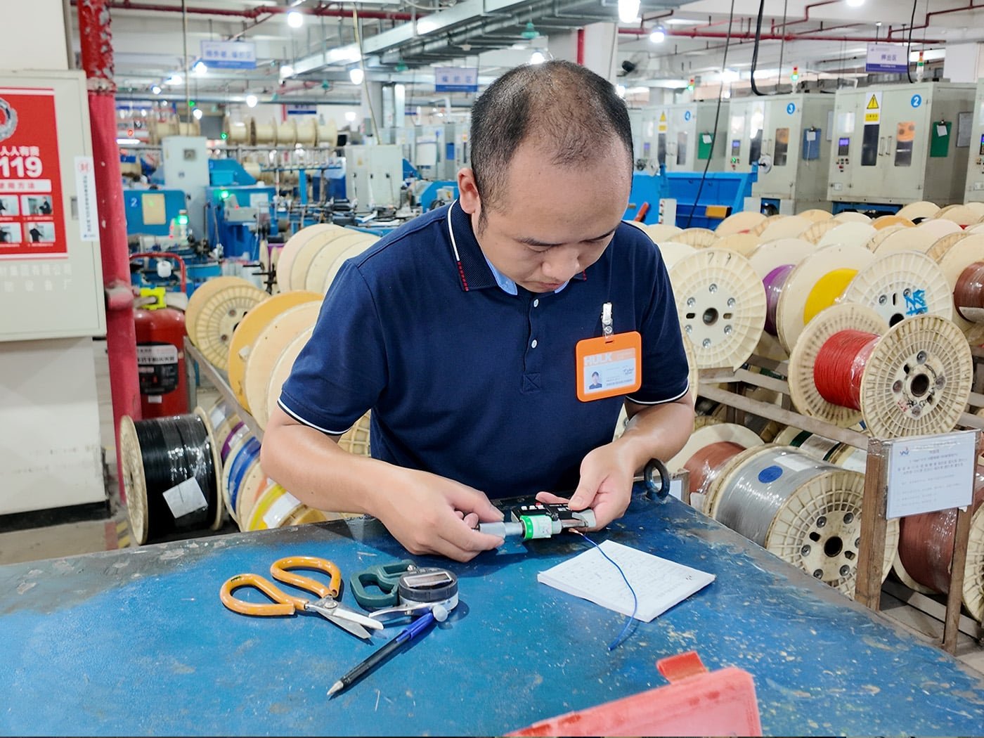 Hulk Cable technician performing precision measurement on a blue cable using a digital micrometer at the production line workstation, surrounded by various colored cable reels, ensuring strict adherence to dimensional specifications and quality standards