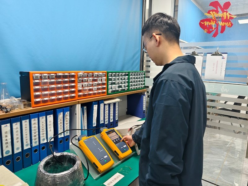 Hulk Cable engineer operating yellow handheld digital testers in a quality control lab, with organized orange and green component bins and blue labeled binders in the background, emphasizing precision measurement and traceable documentation for cable performance validation