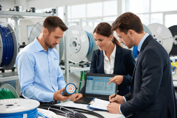 Three engineers in high-visibility vests and hard hats examining an LSZH cable cross-section and reviewing technical data on a laptop in a cable manufacturing facility, illustrating collaborative specification work