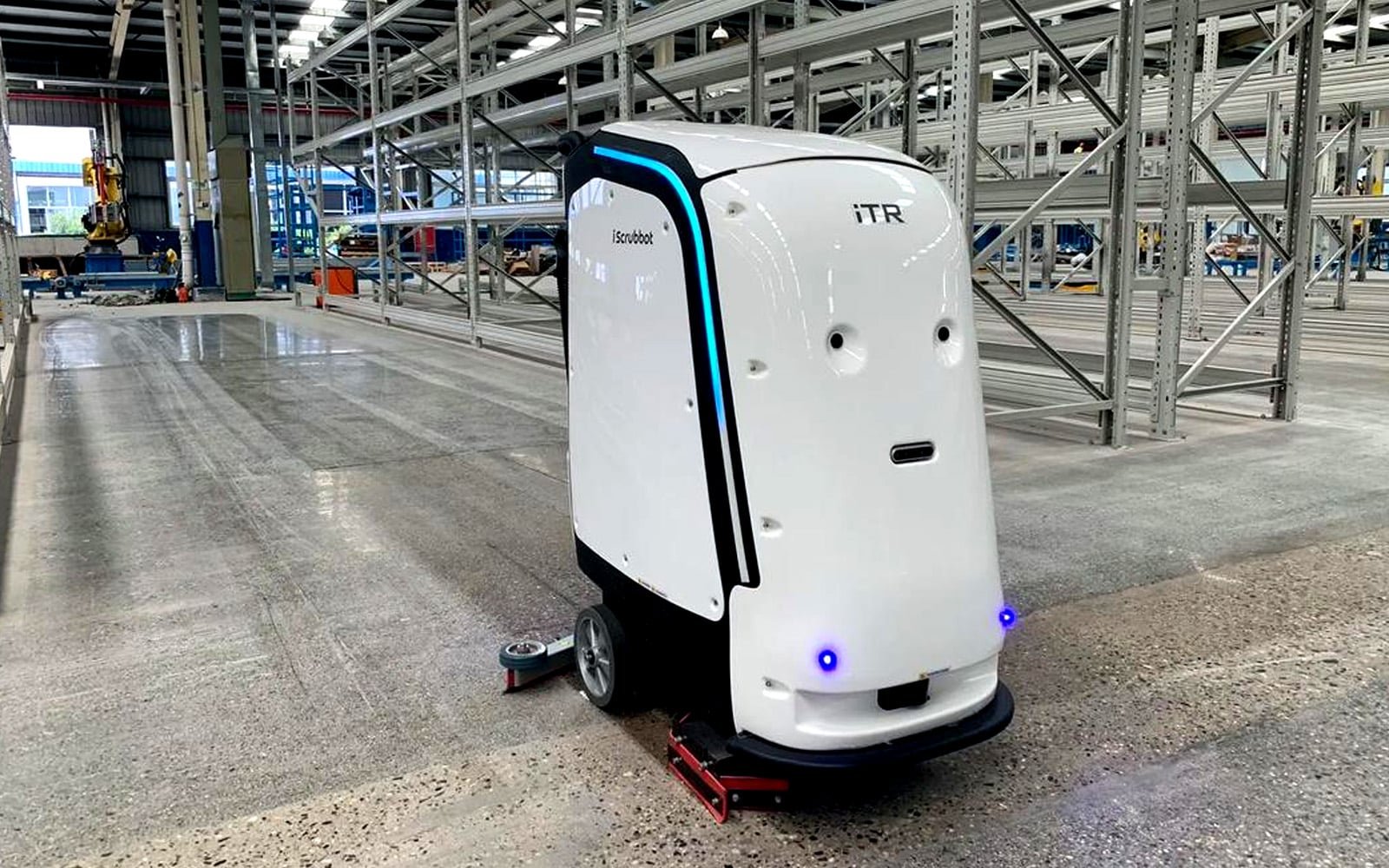 The iScrubbot M2Pro autonomous floor scrubber operating in a large industrial warehouse, featuring blue LED navigation lights and a sleek white body; the robot demonstrates its capability for self-charging, auto water refilling, and waste discharge — enabling uninterrupted 4-hour cleaning cycles across 2,500 sqm per hour in complex logistics environments.